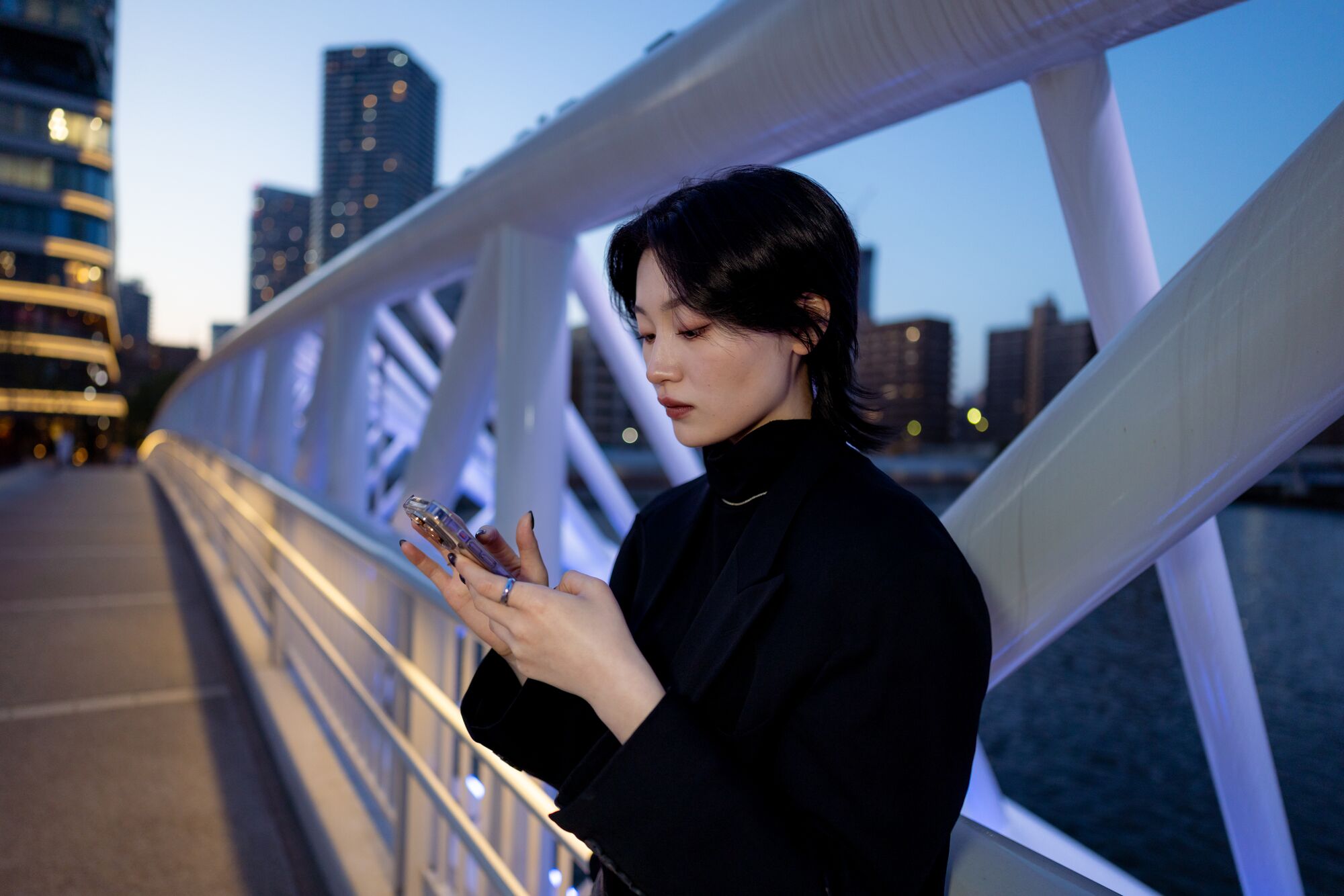 Young woman using smart phone on illuminated bridge in waterfront city in blue hour