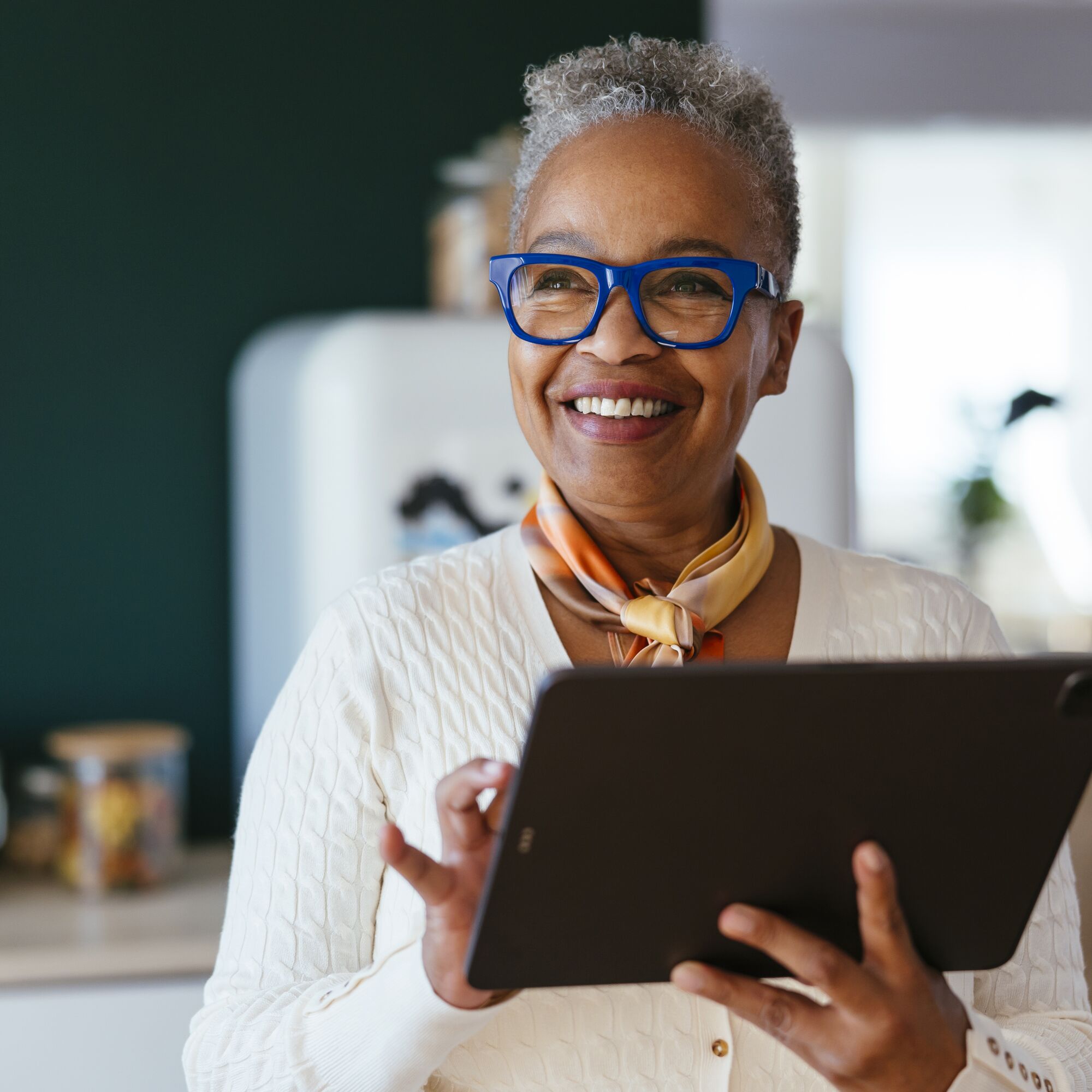 Positive female with grey hair and glasses smiling and looking away while holding a digital tablet in kitchen
