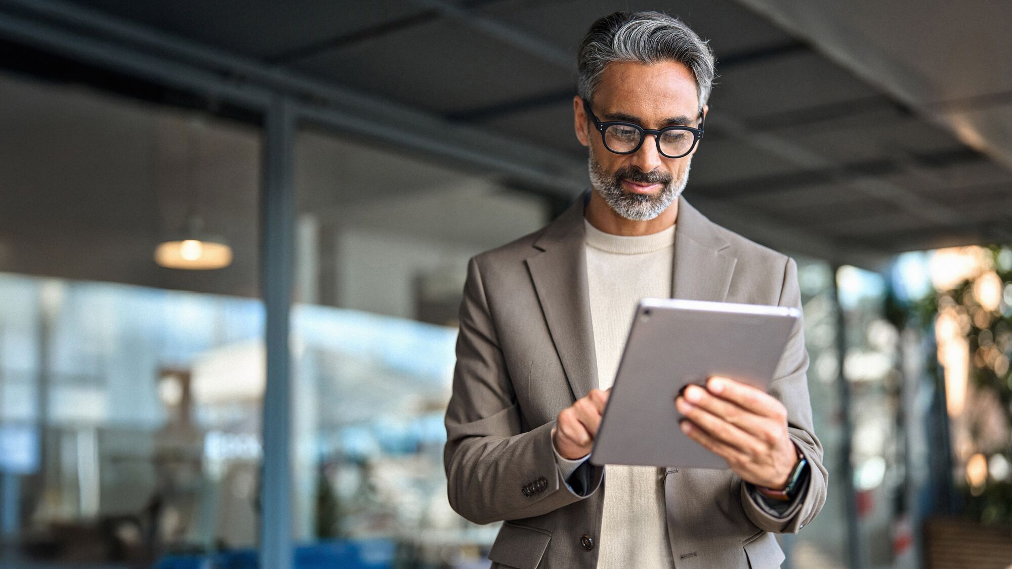 Busy mature older business man executive standing in office using digital tablet. Middle aged professional businessman corporate manager wearing suit and glasses holding tab working on finance project