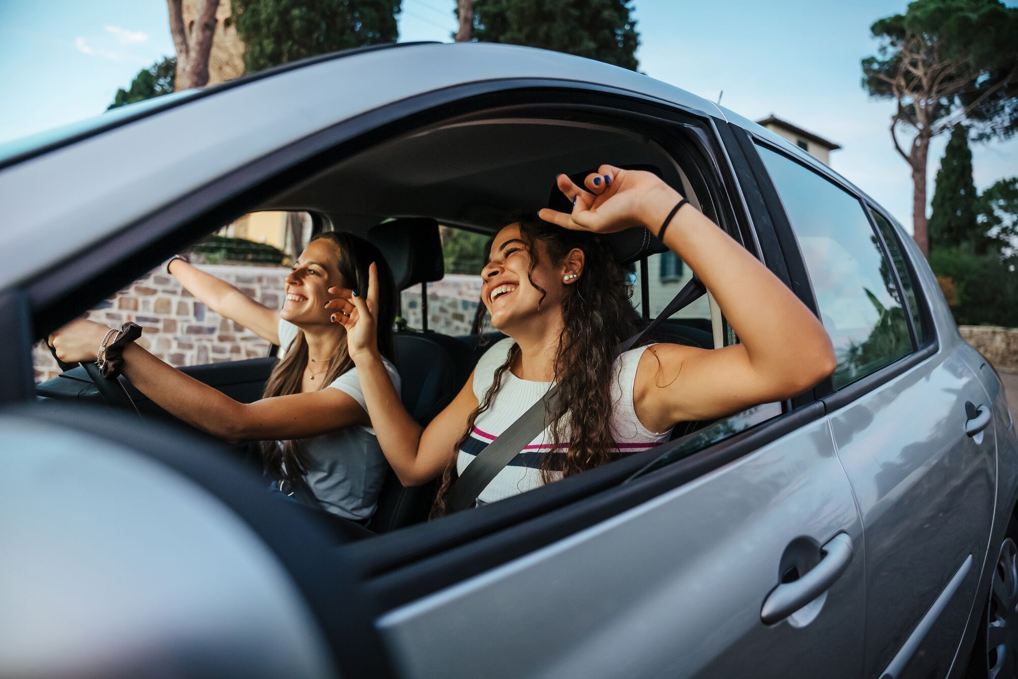 Two young women sing a song on the radio and dance in the car on a day trip in the summer - Best friends having fun together driving around the countryside - Smiling millennial in a relaxing moment