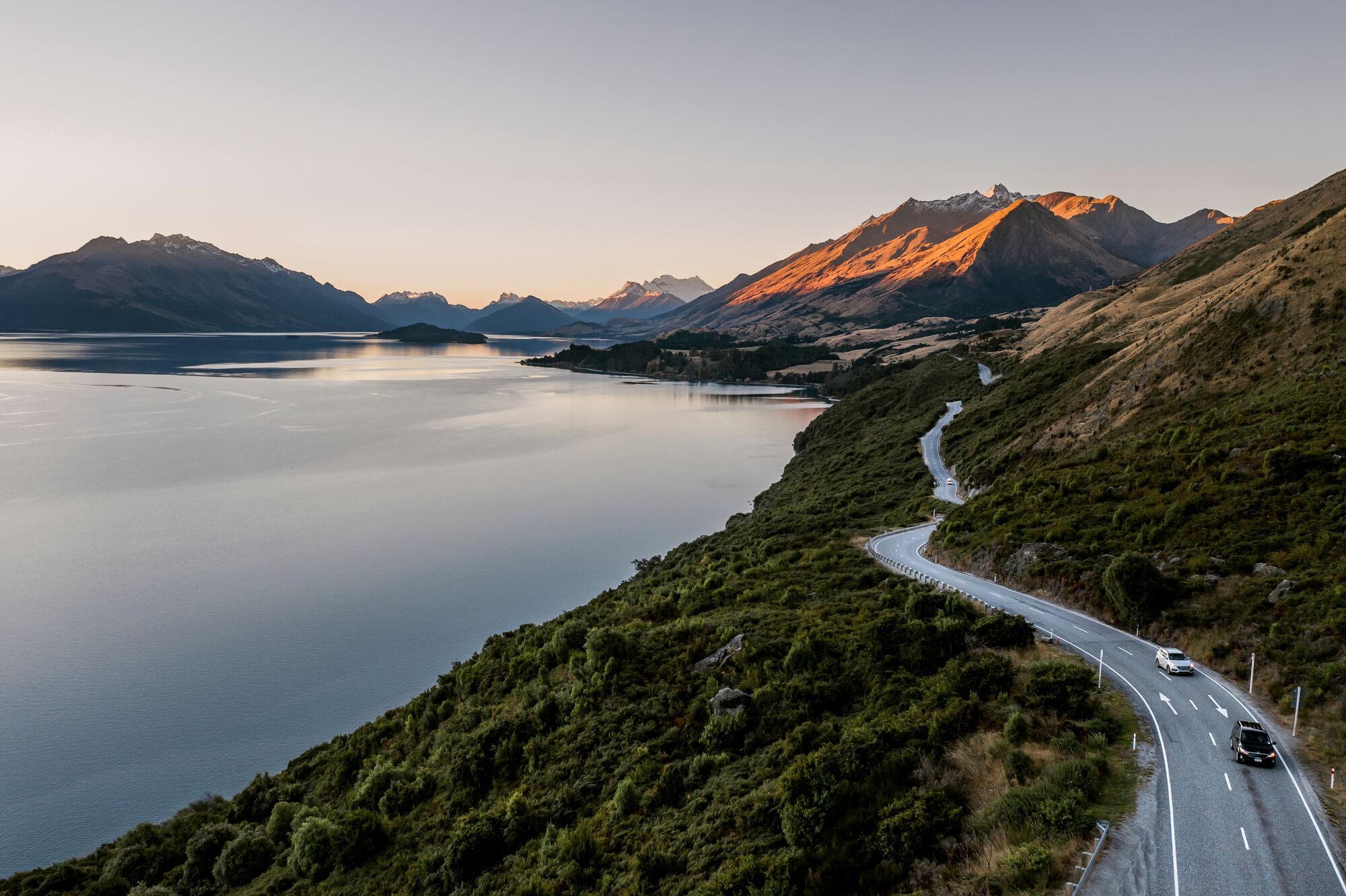 A winding road follows the shoreline of a mountain lake with sunlit peaks in the background.