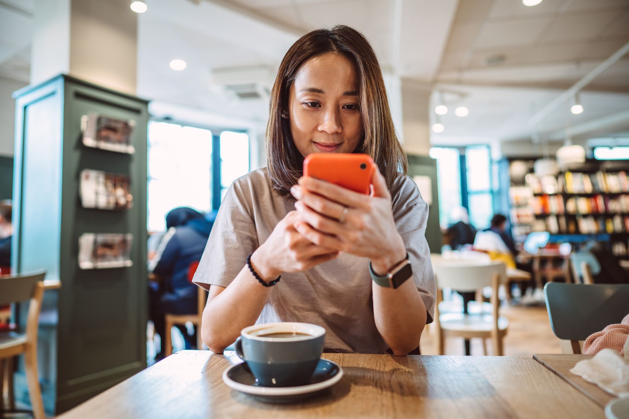 Young pretty Asian woman using smartphone joyfully while enjoying her coffee in a cafe.