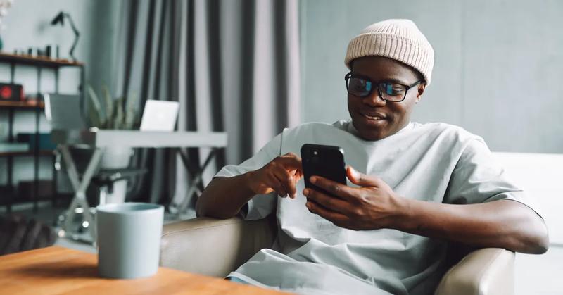 Man wearing glasses and a knit hat smiles while using his smartphone at home.