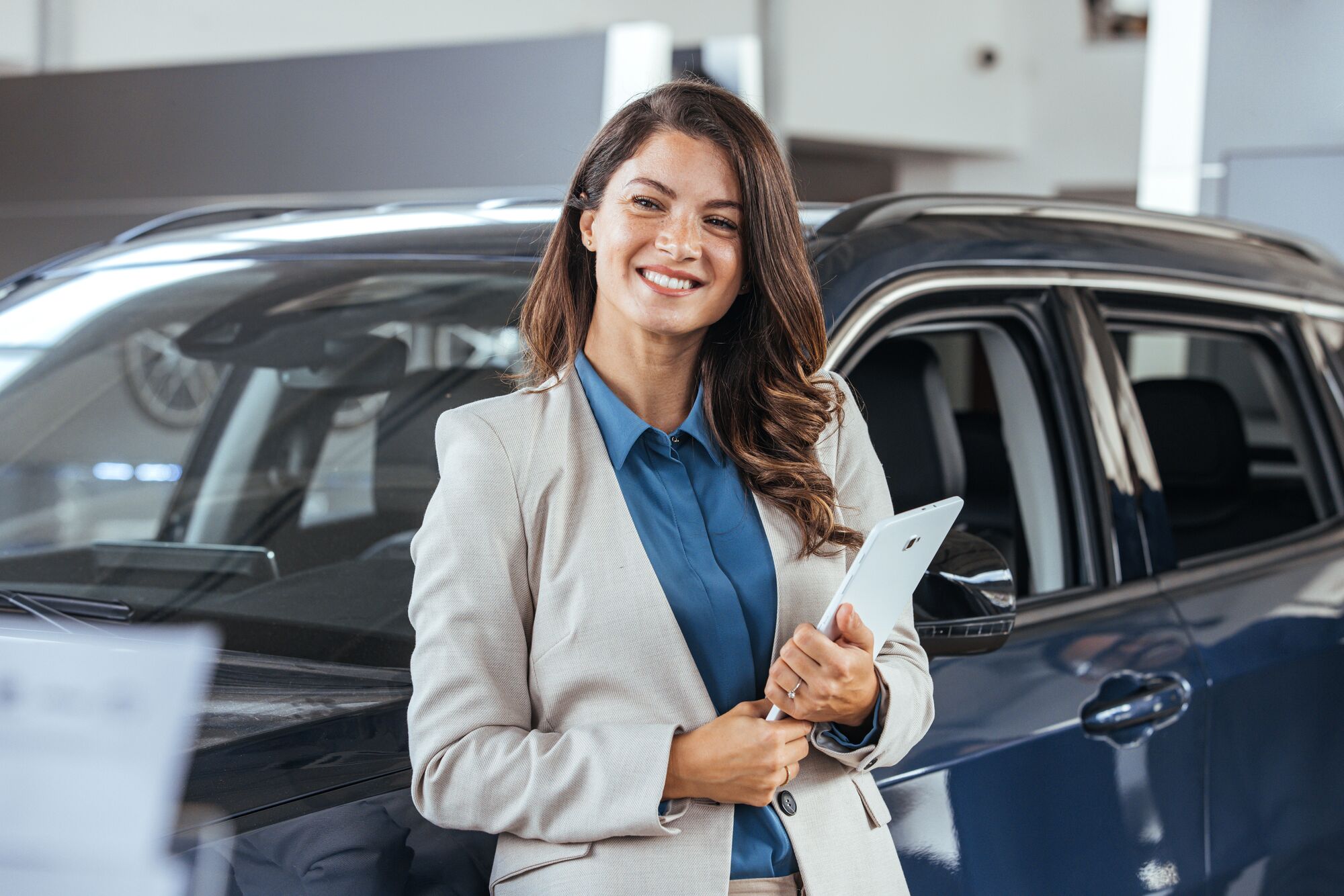 Elegant salesperson working at car dealership. Cheerful young woman with tablet PC smiling and looking at camera while working in modern car dealership. Saleswoman in car showroom selling cars