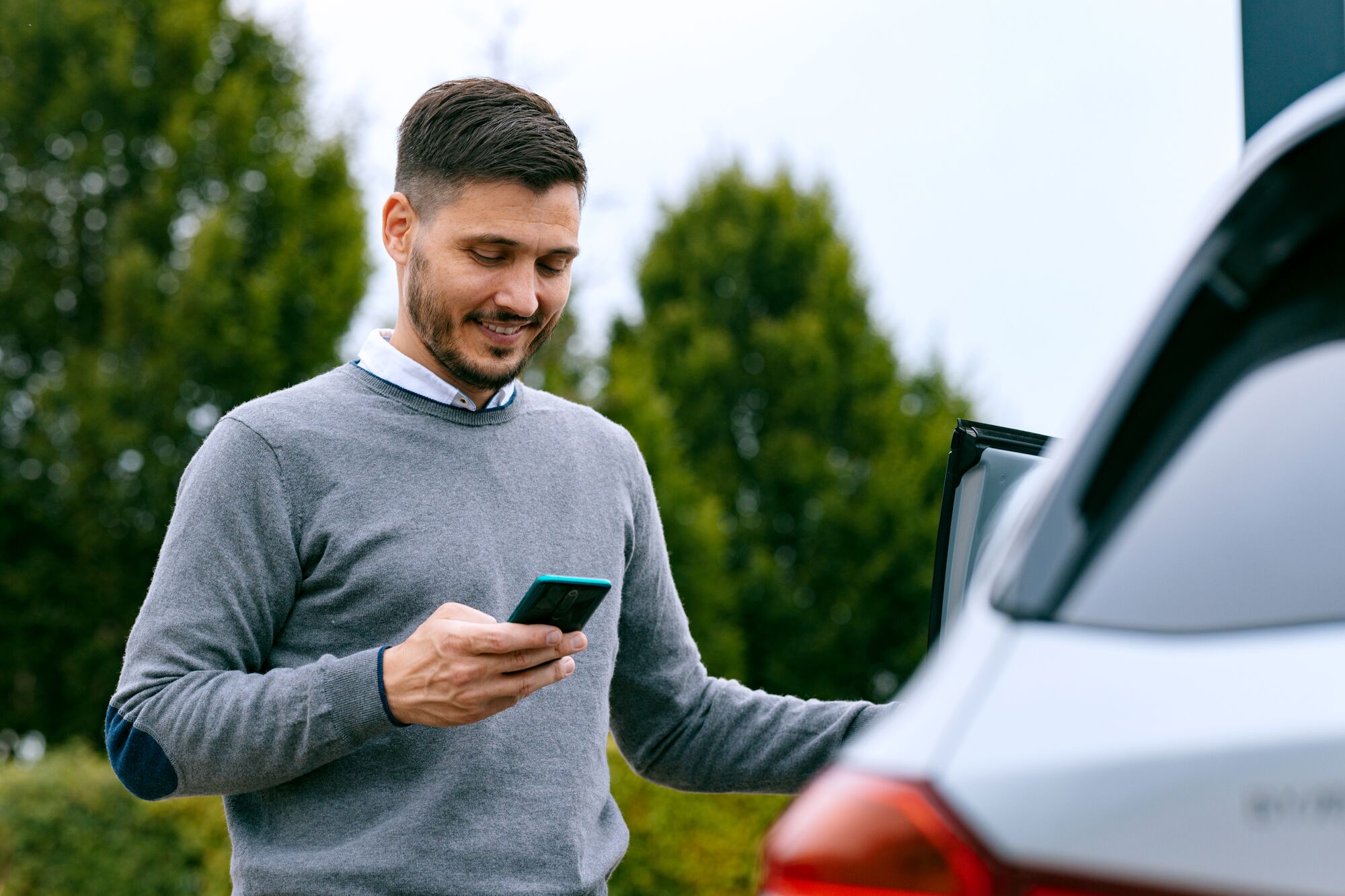 A smiling Caucasian Hispanic male using his smartphone after driving his vehicle.