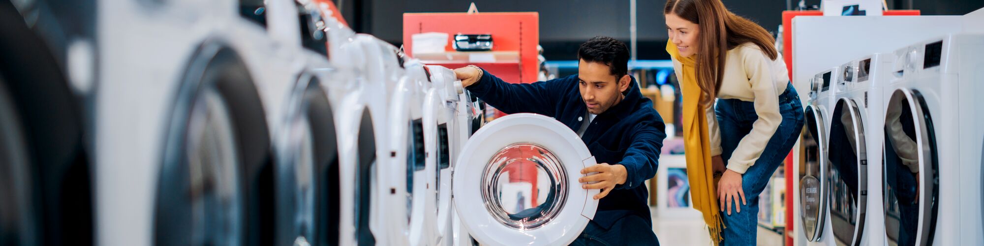 Multicultural Couple Evaluates Washing Machine Choices at Home Electronics Store. Man and Woman in Search of a Reliable Laundry Appliance. Customers Explore Modern Laundry Solutions in Retail Shop