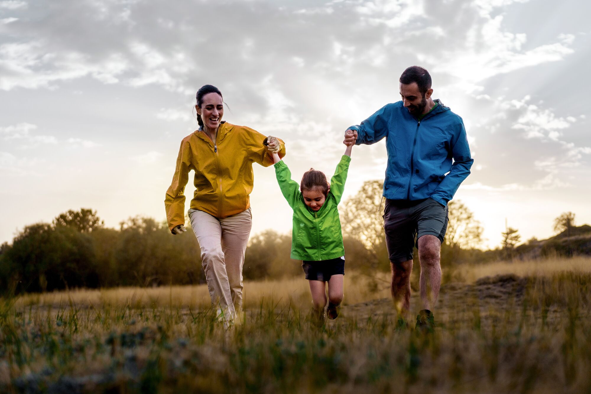 Family enjoys a playful run in the field at sunset, with a child swinging between parents, embodying the joy of active family life and the beauty of nature.
