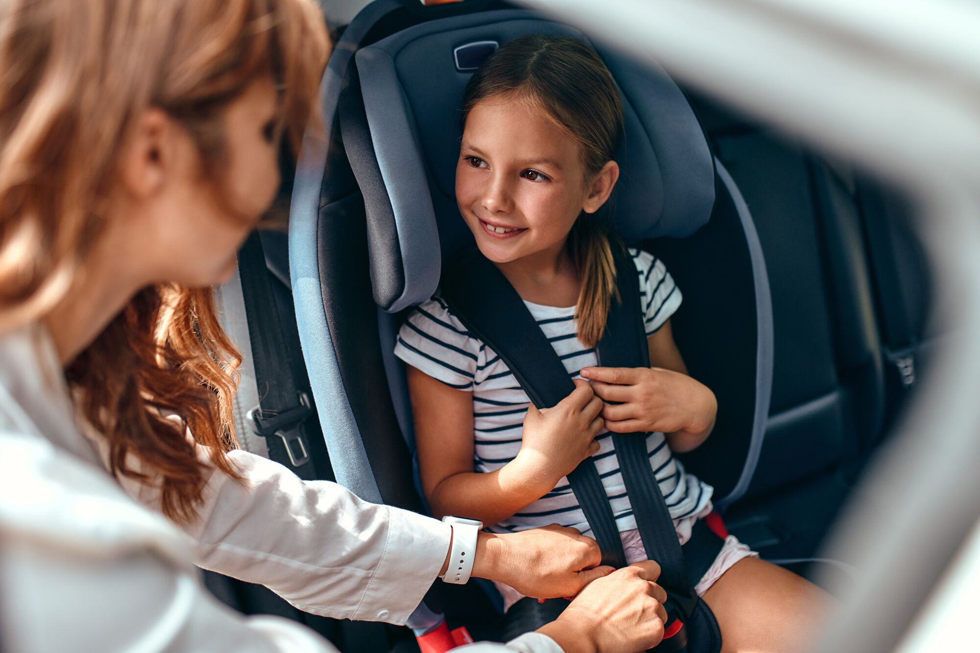 A cute woman mother put her daughter in a car seat and fastens her seat belts. Protection during the trip in the car.