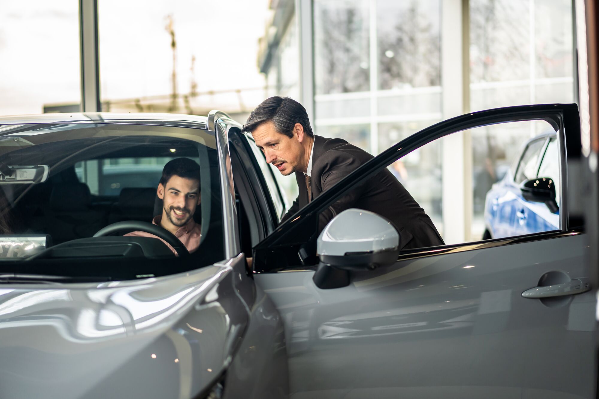 Car salesman consulting smiling customer seated in a new vehicle inside a modern dealership showroom, discussing purchase options and service