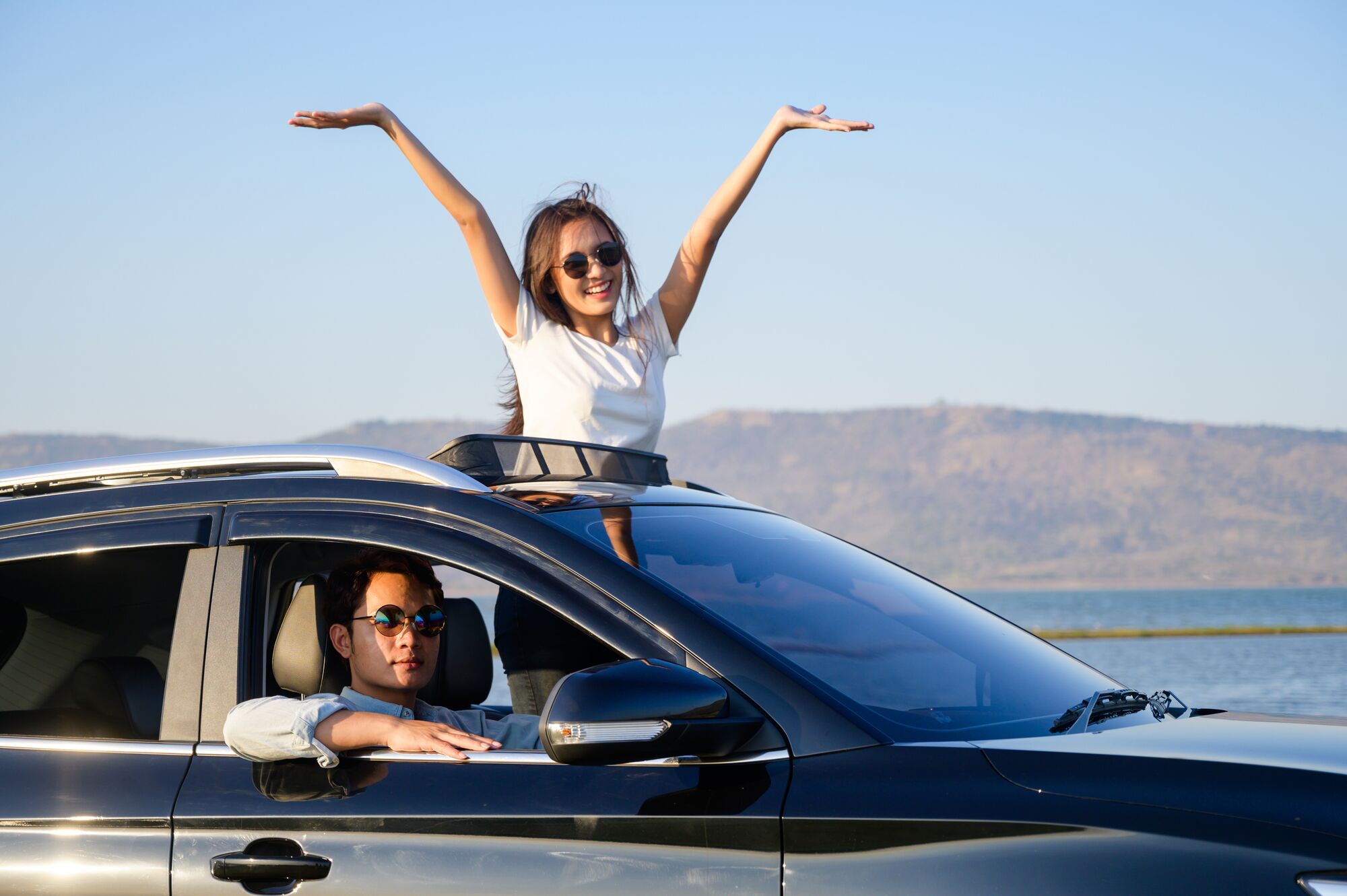 Asian woman in car with raised hands driving on sunset. Front view of young caucasian woman with arms outstretched standing out of sunroof of car with man driving