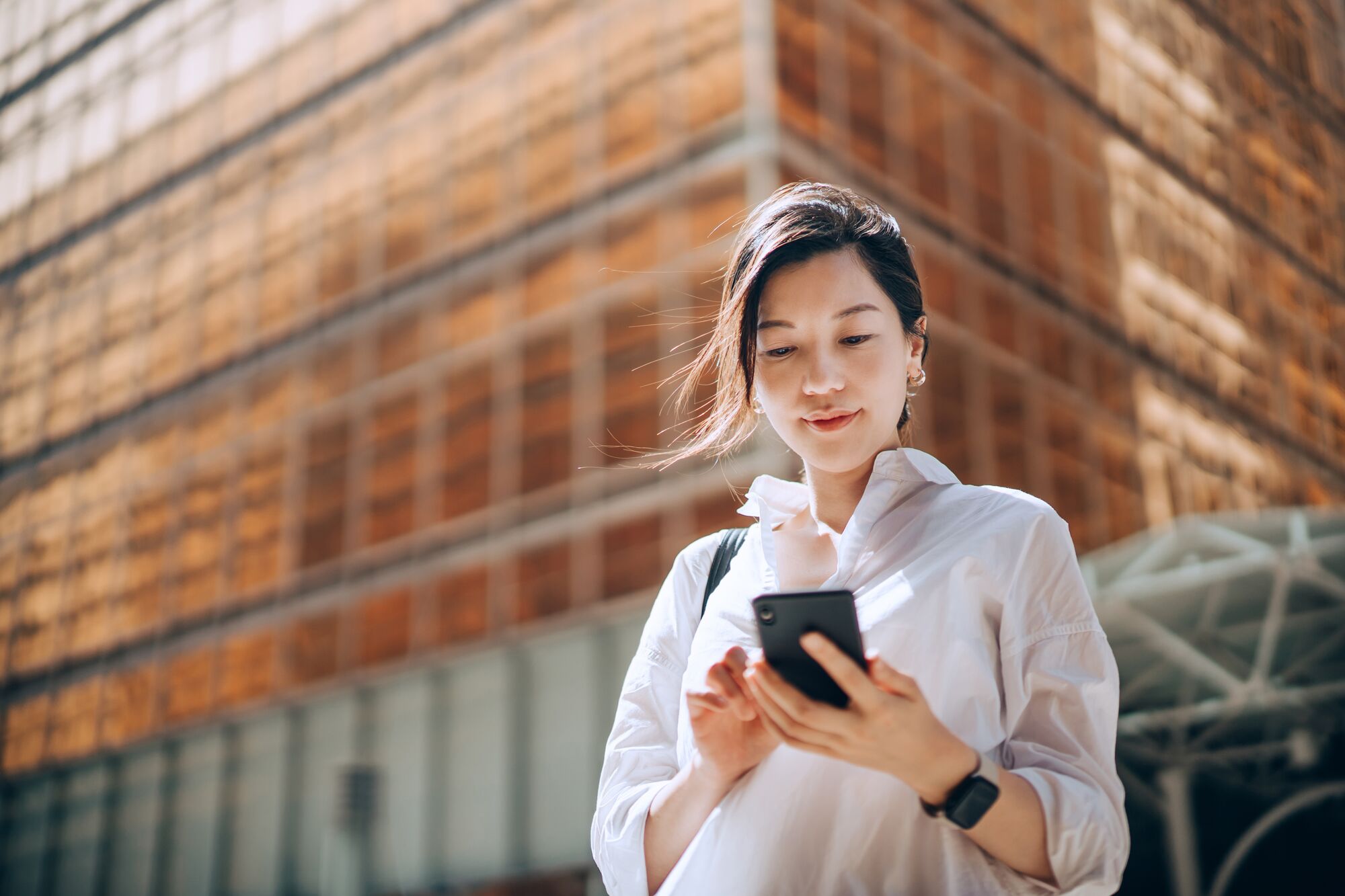 Low angle portrait of young Asian businesswoman checking emails on smartphone outside office building in financial district.