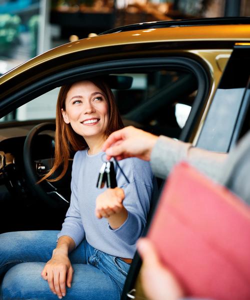 Happy young woman sitting in a new car and receiving keys from a salesman during vehicle purchase at modern car dealership