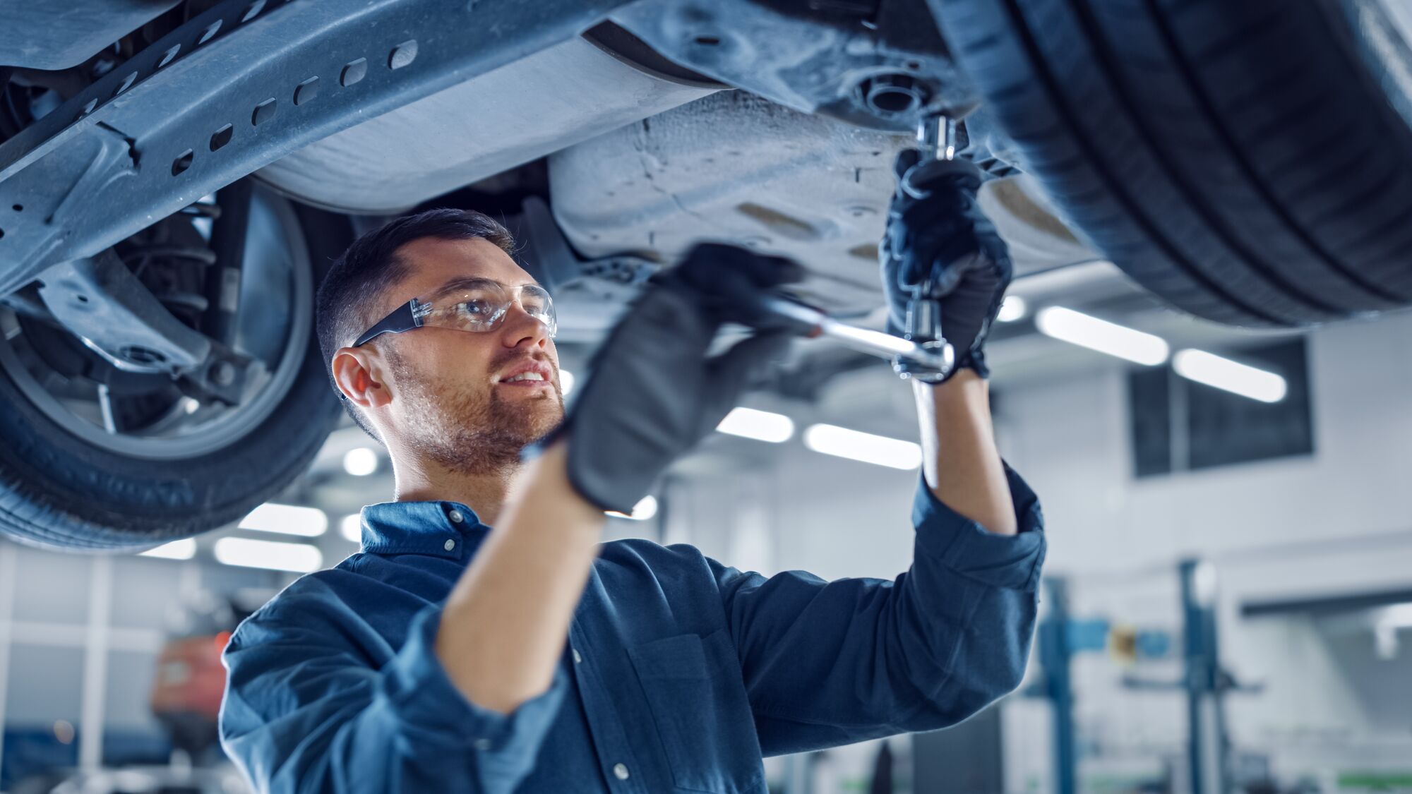 Portrait Shot of a Handsome Mechanic Working on a Vehicle in a Car Service. Professional Repairman is Wearing Gloves and Using a Ratchet Underneath the Car. Modern Clean Workshop.