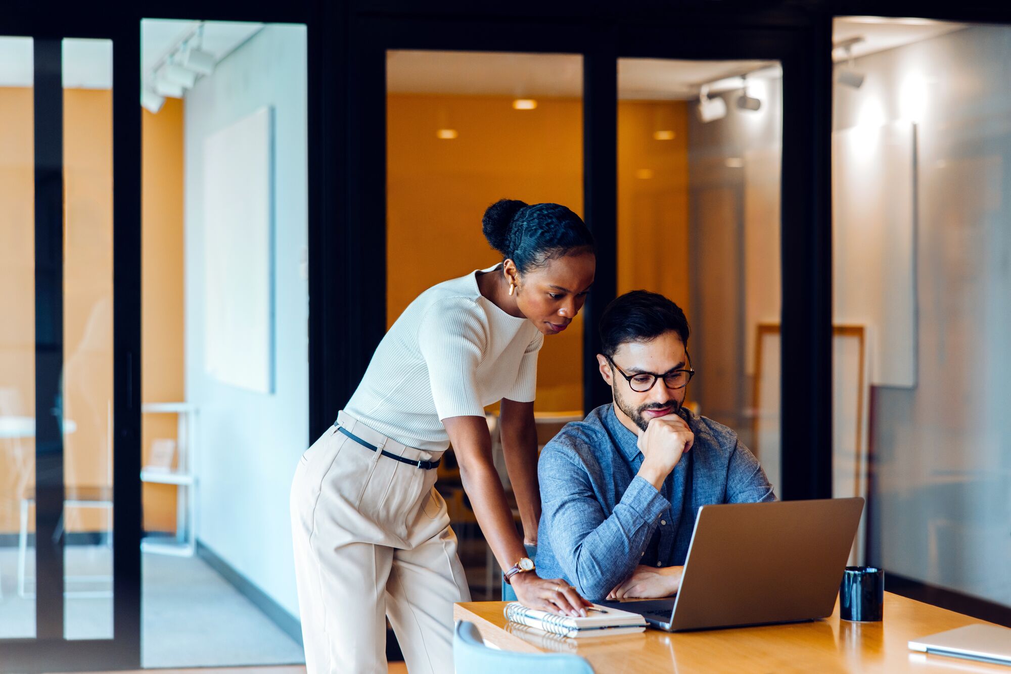 A man sitting and a woman standing reviewing information on a laptop in an office. They are focused while discussing the material, underlining teamwork and effective communication between colleagues.