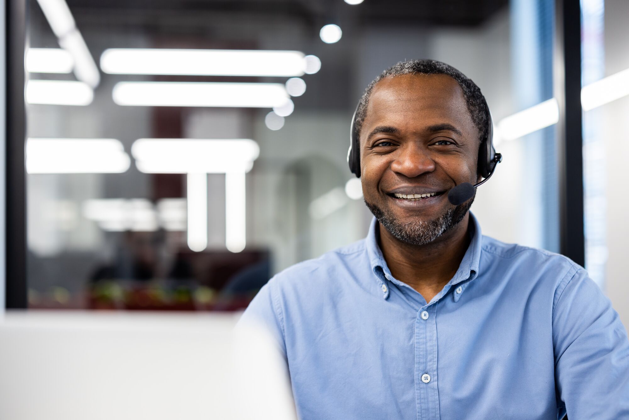 Smiling customer support agent wearing headset working in office. Professional expression conveys positivity and readiness to assist. Modern office setting enhances professional appearance