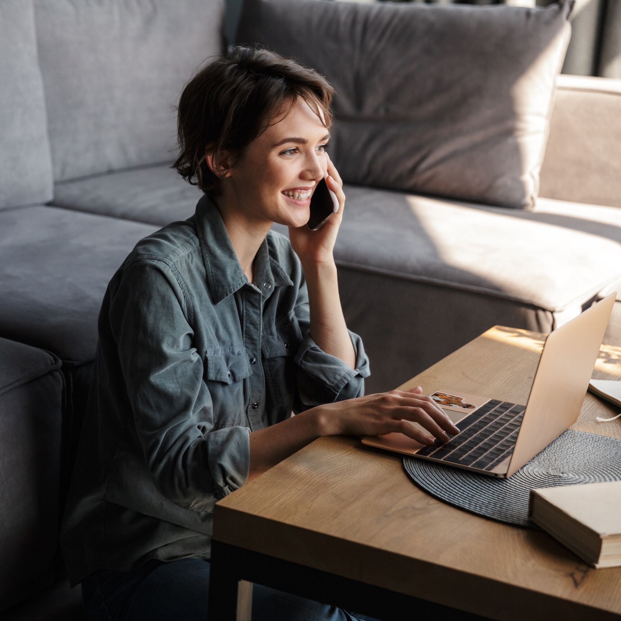 Image of young cheerful woman using laptop and talking on cellphone while sitting on floor at living room