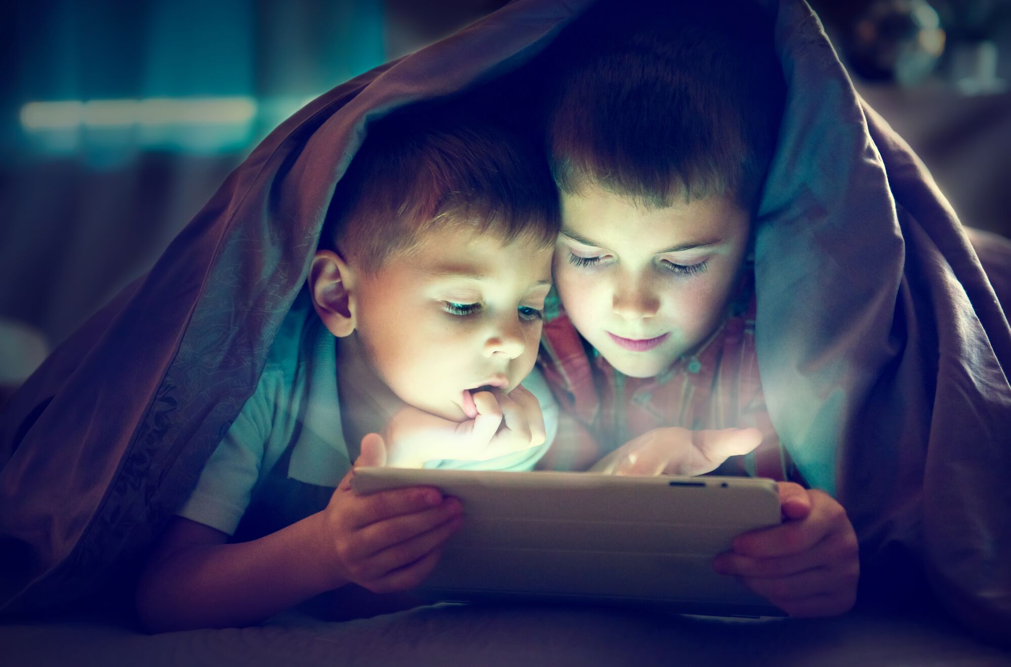 Two kids using tablet pc under blanket at night.