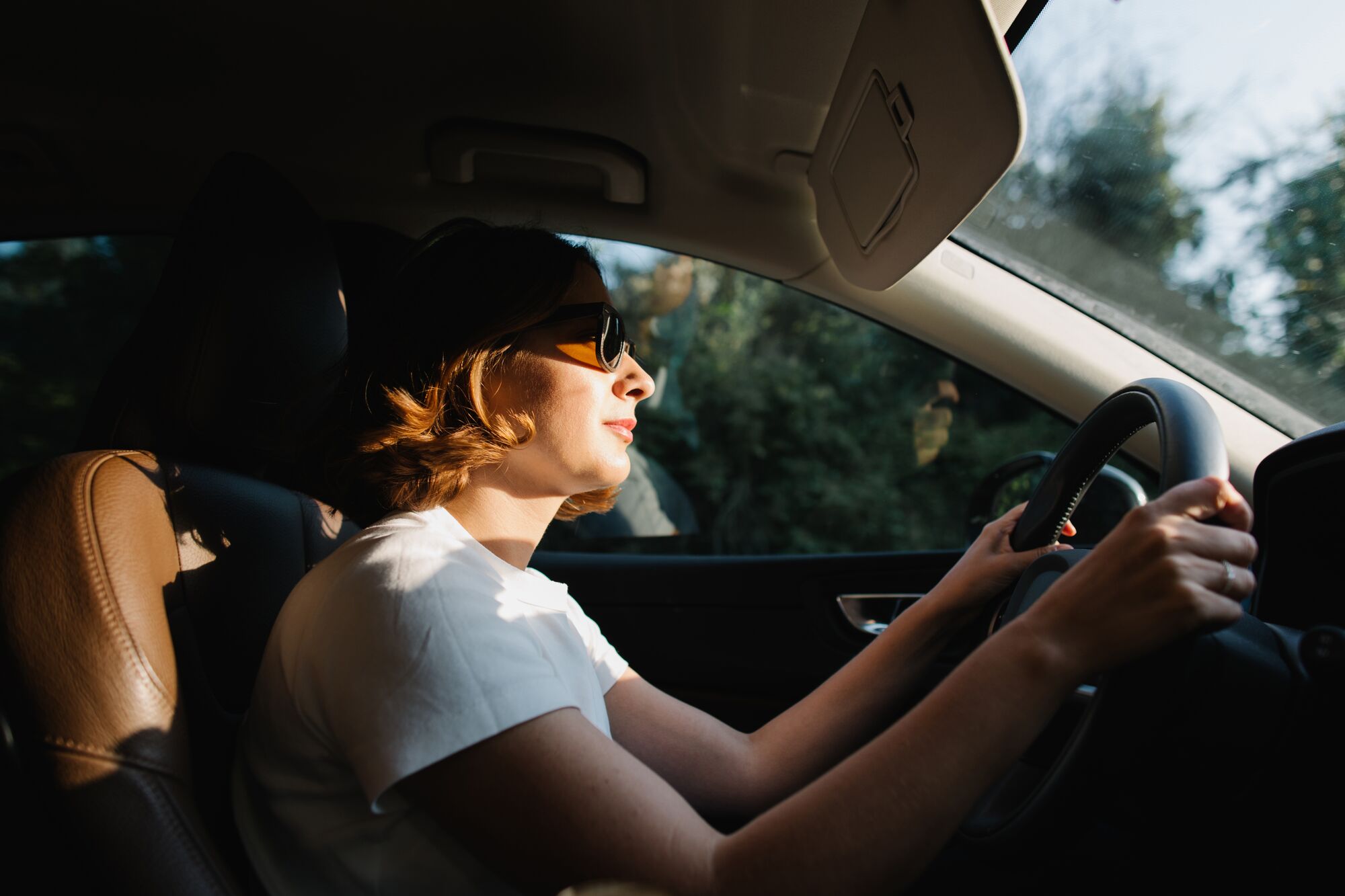 A woman in sunglasses with short haircut driving a car in the forest with the sunset light