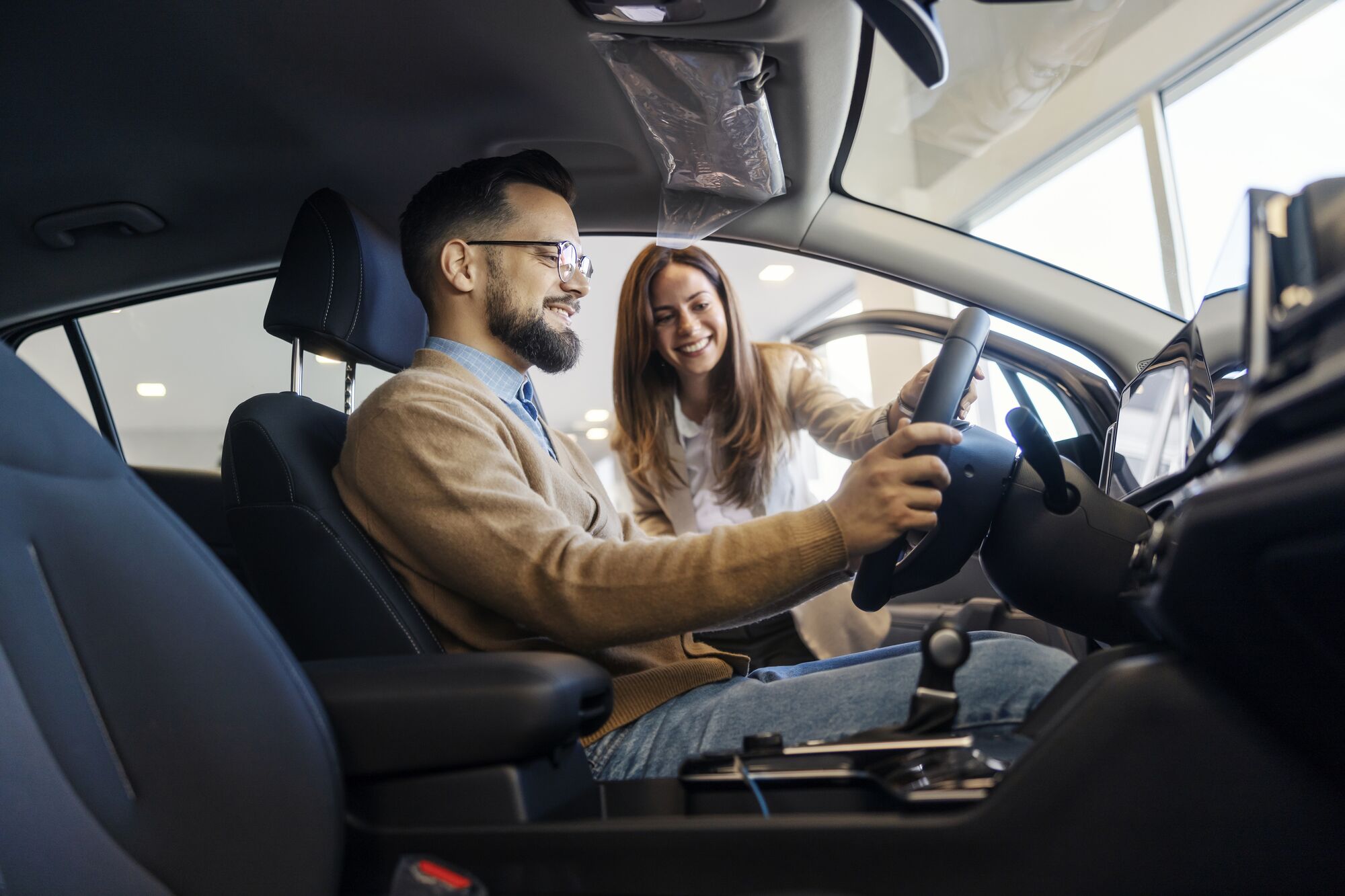 Side view of smiling buyer sitting in luxury car while female car seller showing performances.